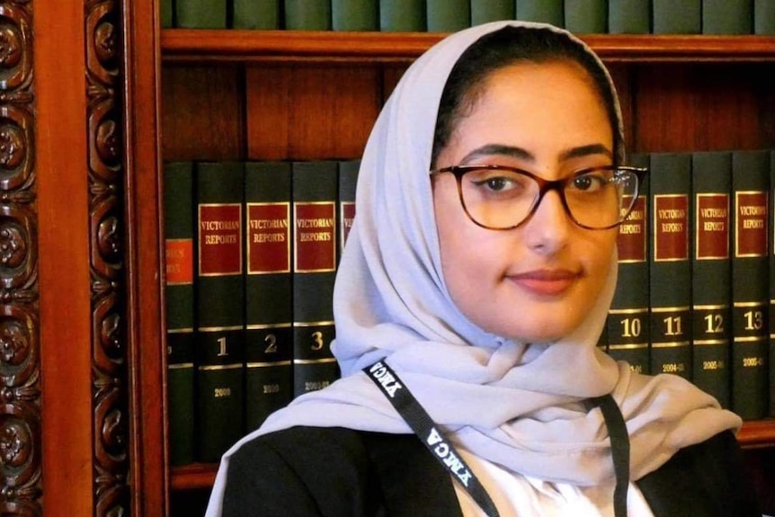 A young woman looks into the camera while wearing a lilac headscarf and sitting before a shelf of bound leather books.