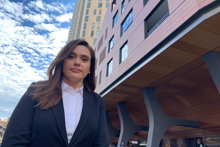 Une femme en blazer foncé et chemisier blanc se tient devant un immeuble de bureaux.