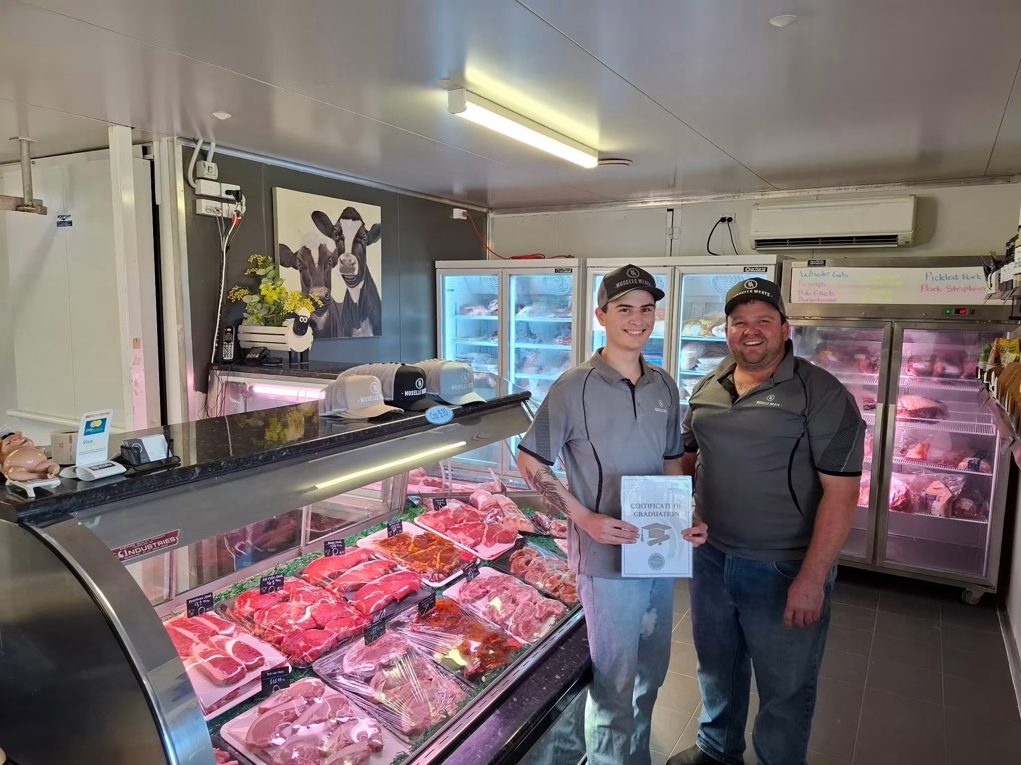 A young man with an older man standing in a butcher shop in front of the meat case, smiling looking into the camera.