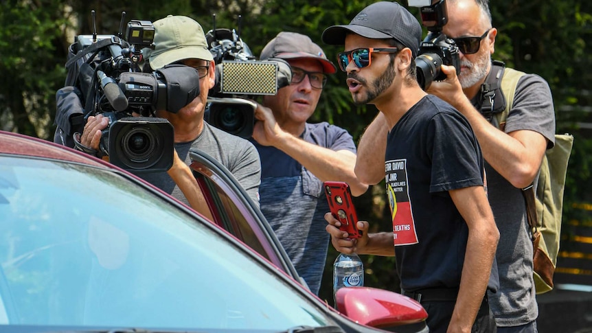 A man appears to shout at a person in a car as news cameras film the interaction.