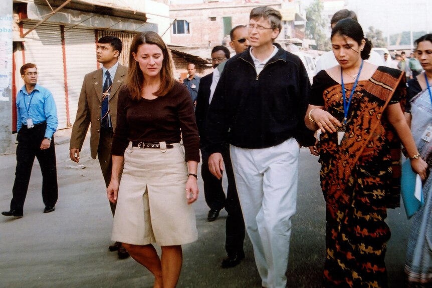 Melinda and Bill Gates walk on a street surrounded by people with lanyards. 