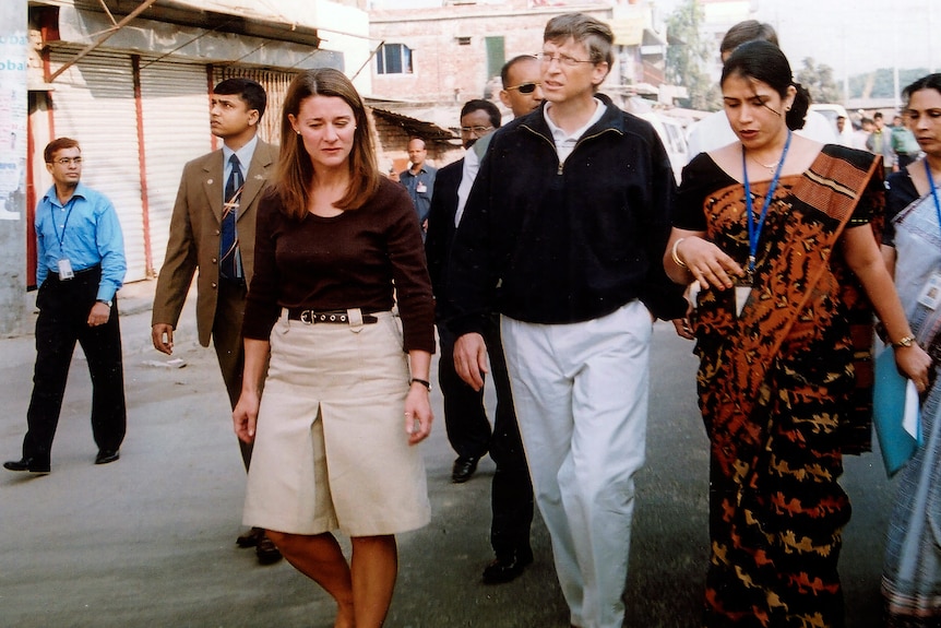 Melinda and Bill Gates walk on a street surrounded by people with lanyards. 