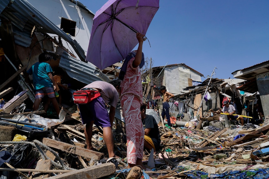A woman holds a parasol over two people who are looking through the wreckage of home brought down in a typhoon.
