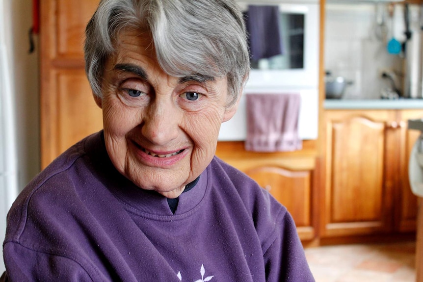 An older woman with grey hair, wearing a purple top, sitting in her kitchen.