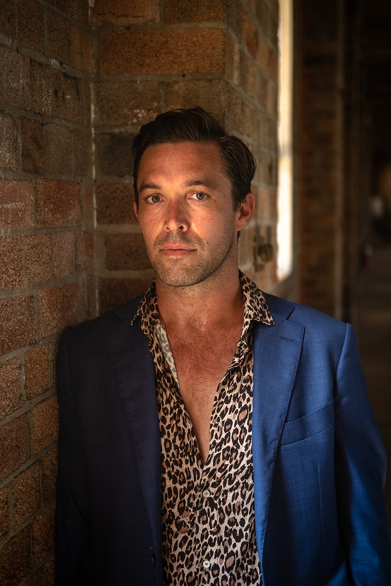 A 30-something Aboriginal man stands in front of a brick wall. He is wearing a blue blazer and leopard print button-up shirt.