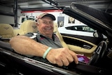 A man in a hat sitting behind the wheel of a car, arm rested on the edge, smiling