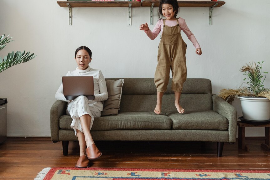 A woman sitting on a couch and typing on a laptop while a young girl jumps up and down next to her