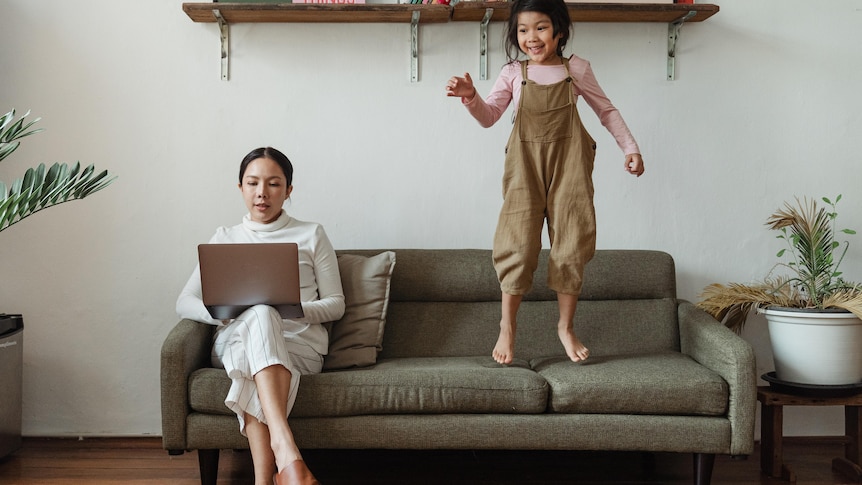 A mother is working on her laptop, while her young daughter jumps on the couch, in story about kids school in lockdown..