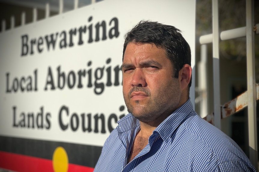 A man with short dark hair and rugged stubble stands in front of an Indigenous land council sign.