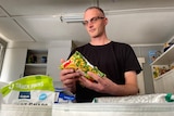 A man waring a black t-shirt and glasses holds a bag of frozen vegetables.