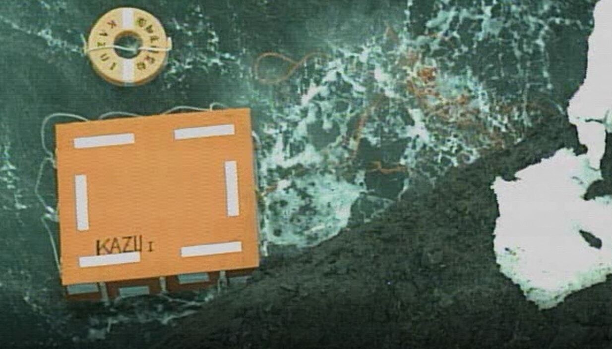 A birds-eye view of floatation devices, a box labelled 'KAZU 1' and some ropes floating in water near rocks.