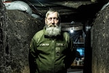 An old bearded man stands in a dug out tunnel, dimly lit by a single lightbulb overhead
