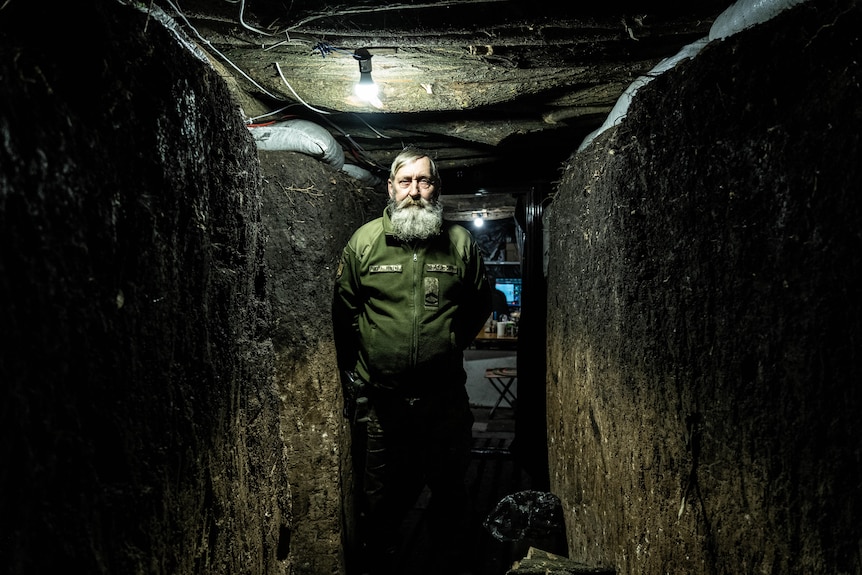 An old bearded man stands in a dug out tunnel, dimly lit by a single lightbulb overhead