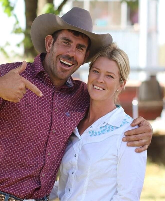 A man in a cowboy hat points at a woman in a white shirt and both are smiling at the camera