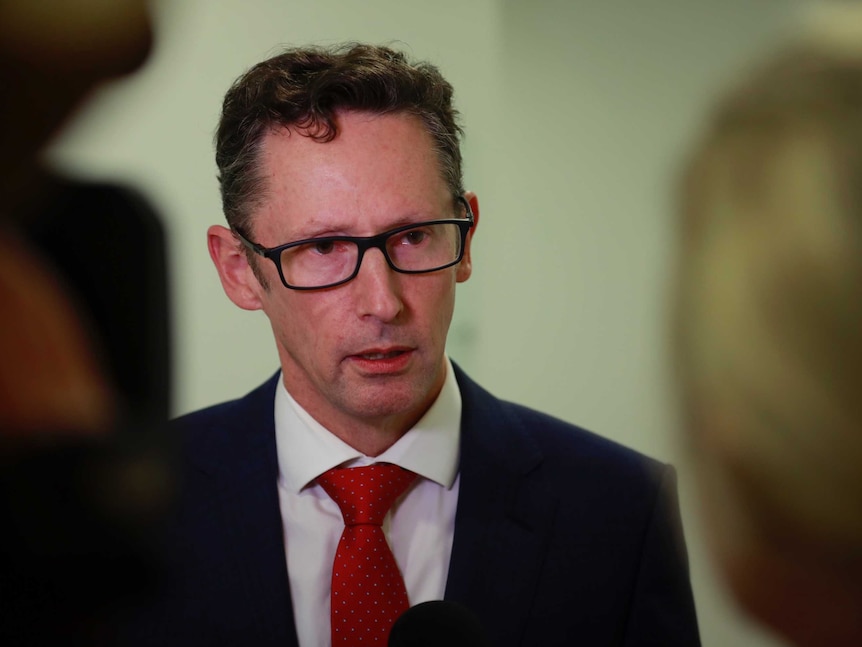 A white man with short brown hair wearing a suit and a red tie speaking to a report who is blurred out in the foreground