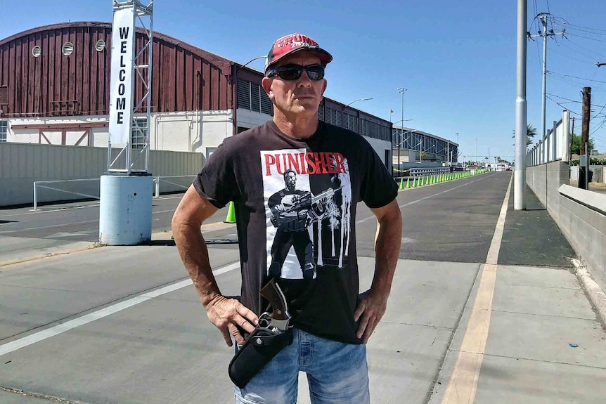 A man with a black hat, black t-shirt, and holstered firearms poses in front of a sports arena