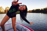 Ebony Forsyth strikes a pose with peace hand symbols while standing on the back of a ski boat on the river.