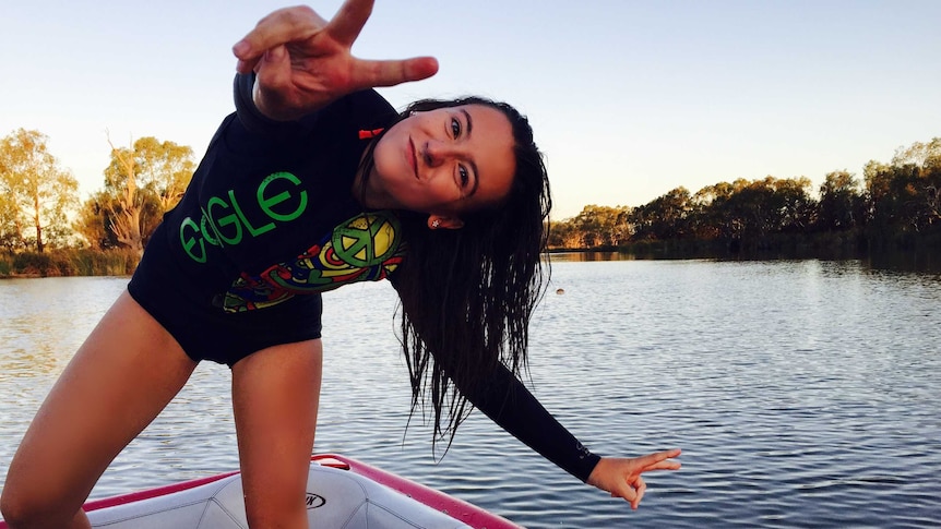 Ebony Forsyth strikes a pose with peace hand symbols while standing on the back of a ski boat on the river.