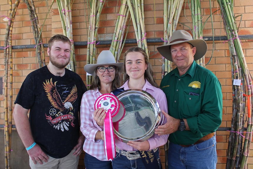 A family of two men, a woman and a teenage girl hold a show ribbon and plate in front of cane stalks.