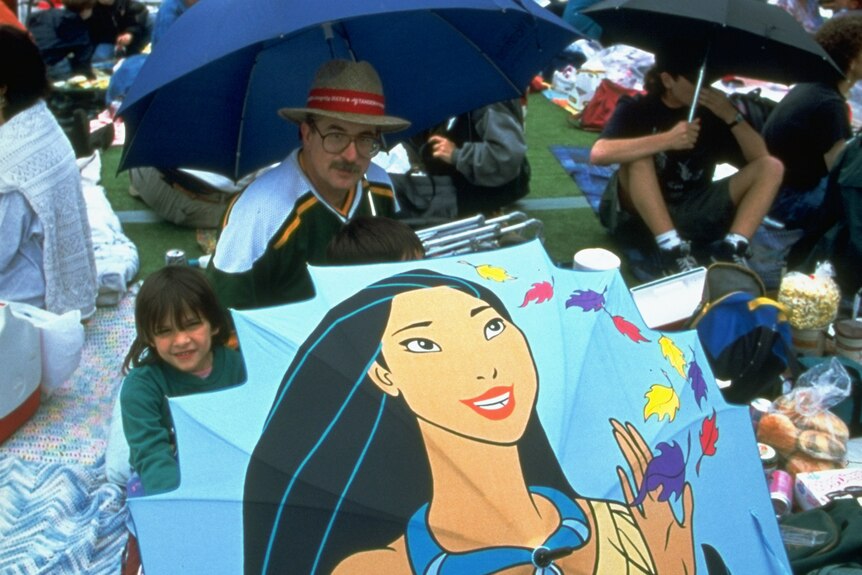 A large crowd of people on grass, with a man holding one large, open umbrella that has cartoon image of Disney's Pocahontas.
