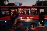 You look at a red, blue and white public bus captured in mid-motion while a person on a scooter blurs past it.