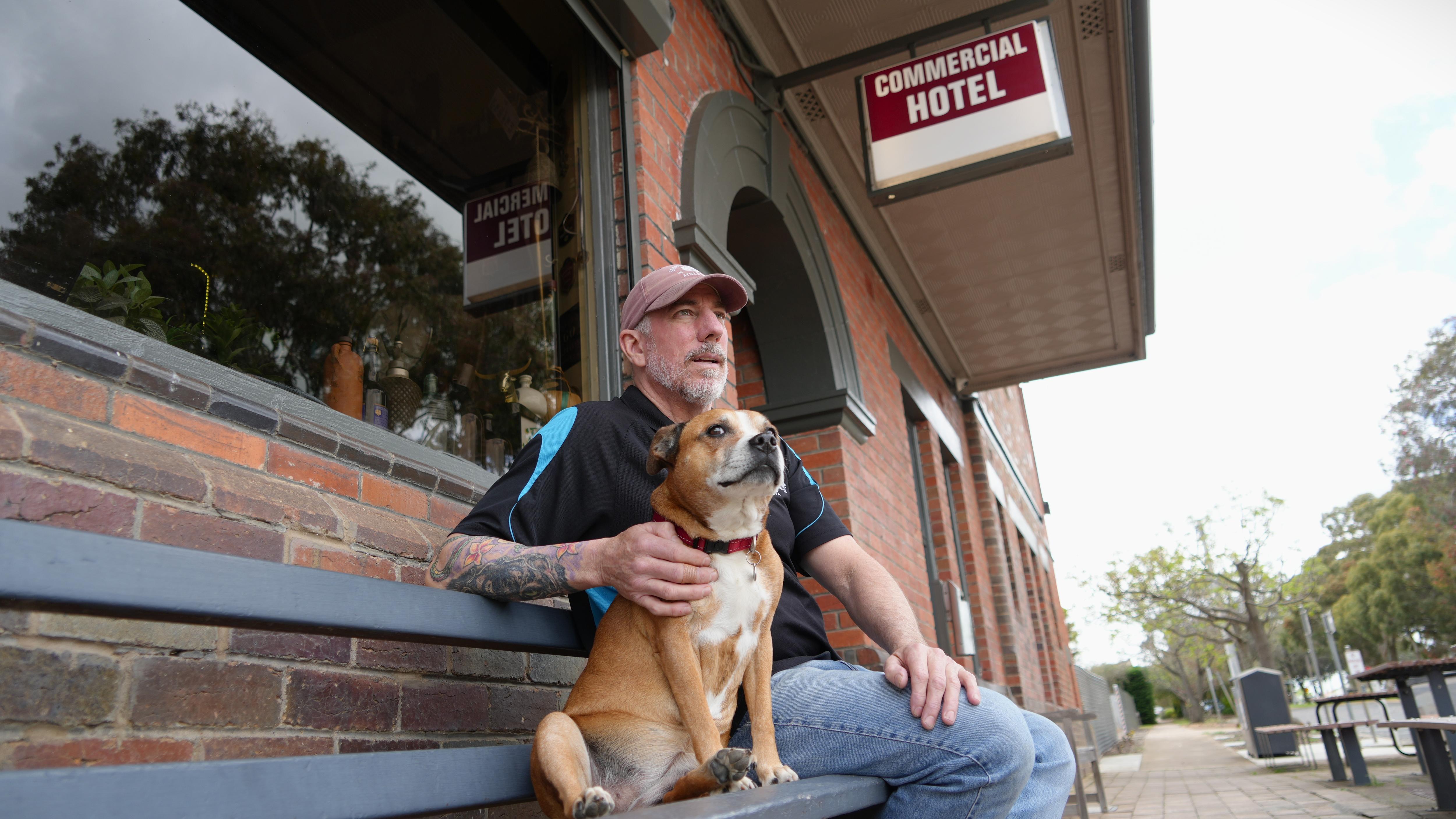 A man and dog sit on a bench outside a building looking towards the town with a sign above them reading: "Commercial Hotel."