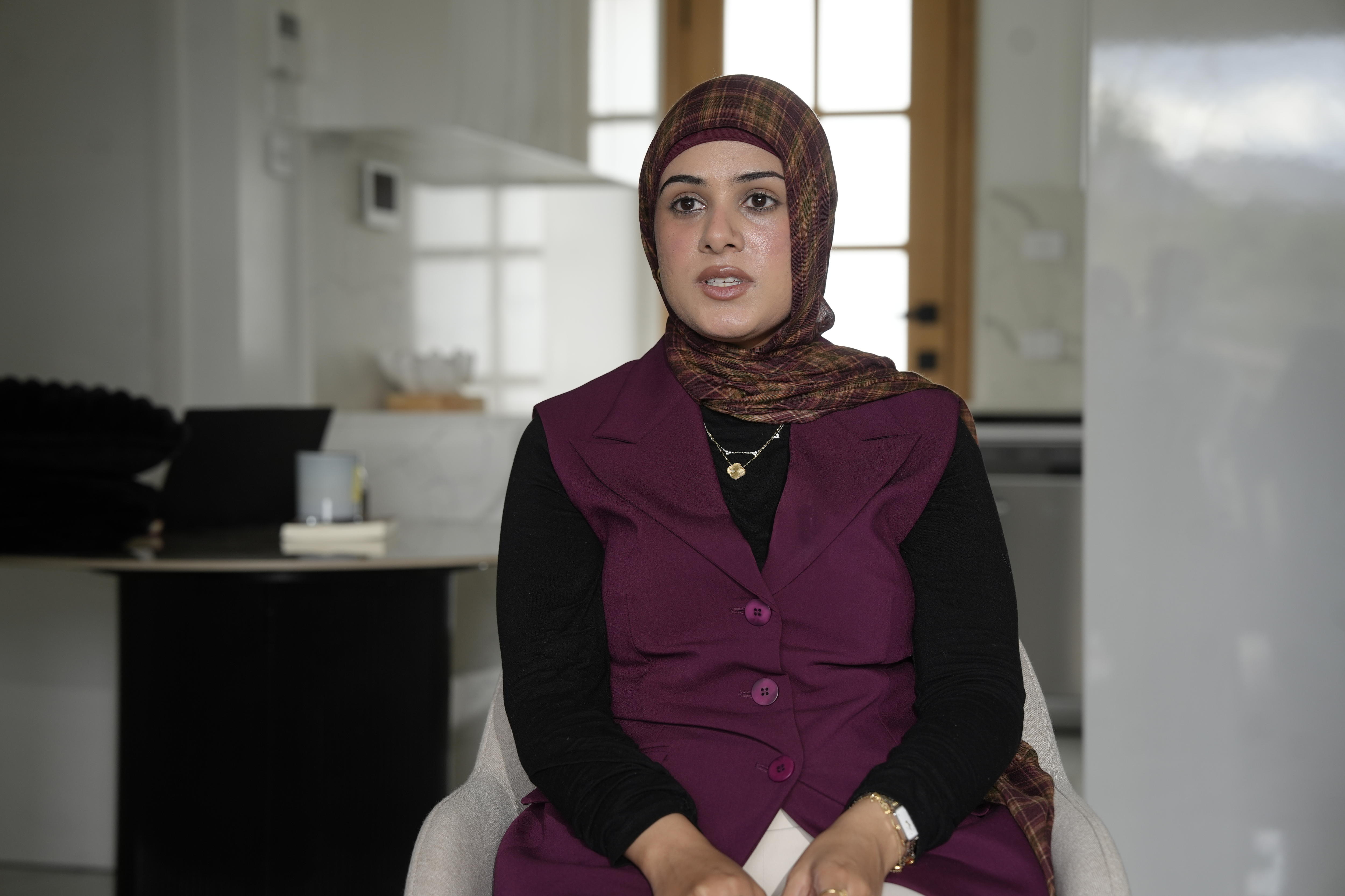 A photo of a woman in a red and brown hijab and red vest with black sleeves, behind her is a office space.