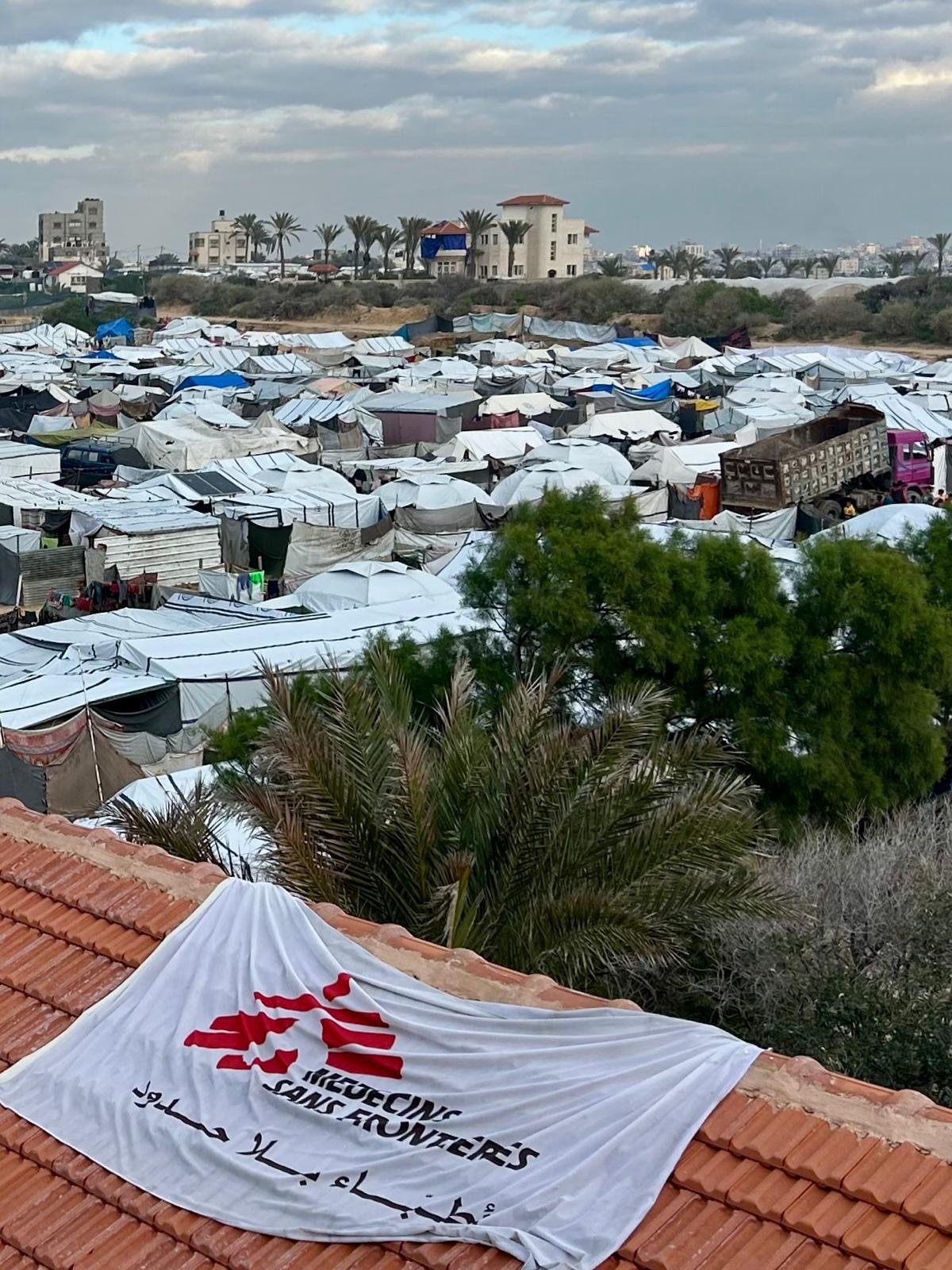 Telhado vermelho com bandeira de MSF e centenas de tendas ao fundo.