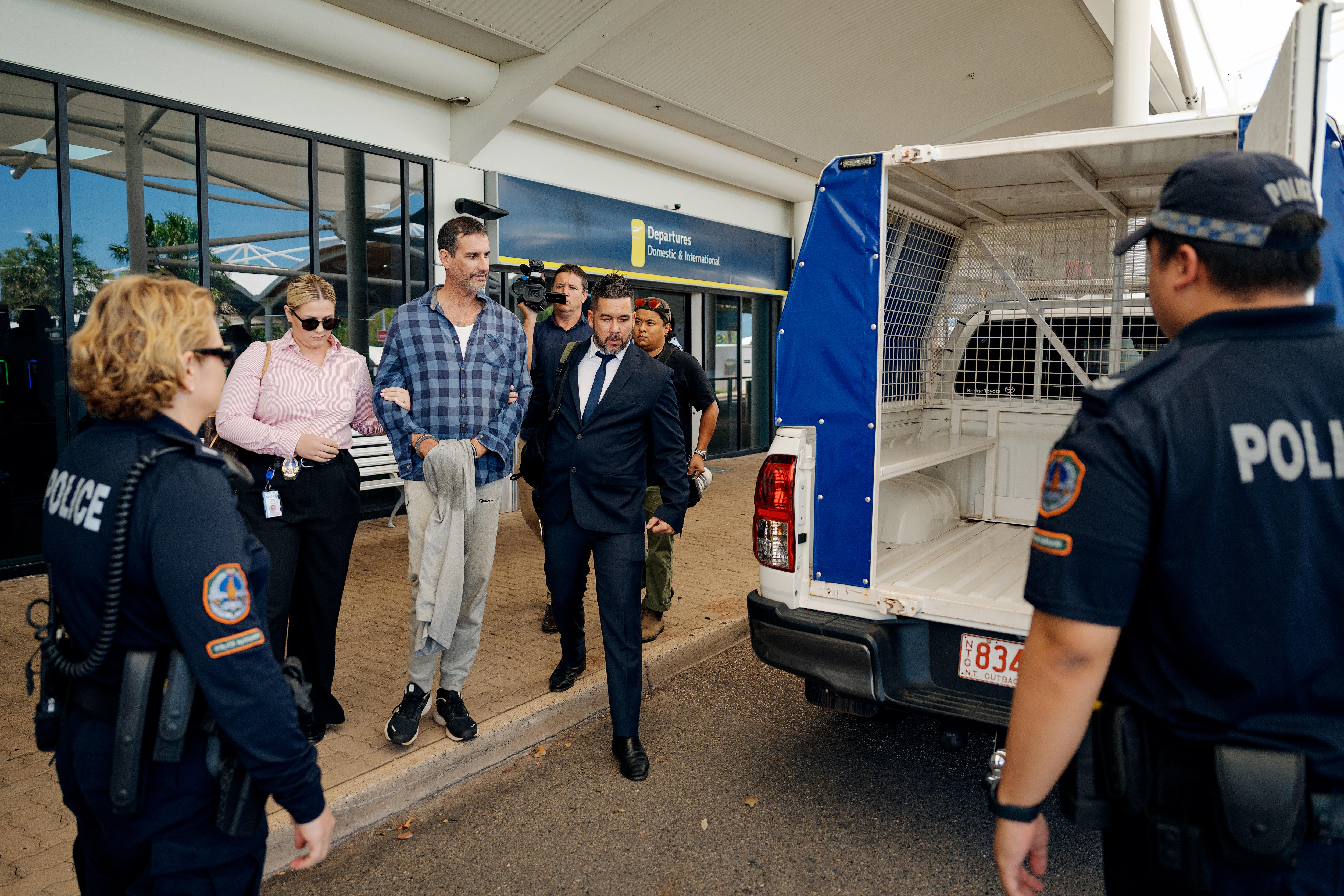 Agentes de policía escoltan a un hombre hasta la parte trasera de un coche de policía frente a un aeropuerto.