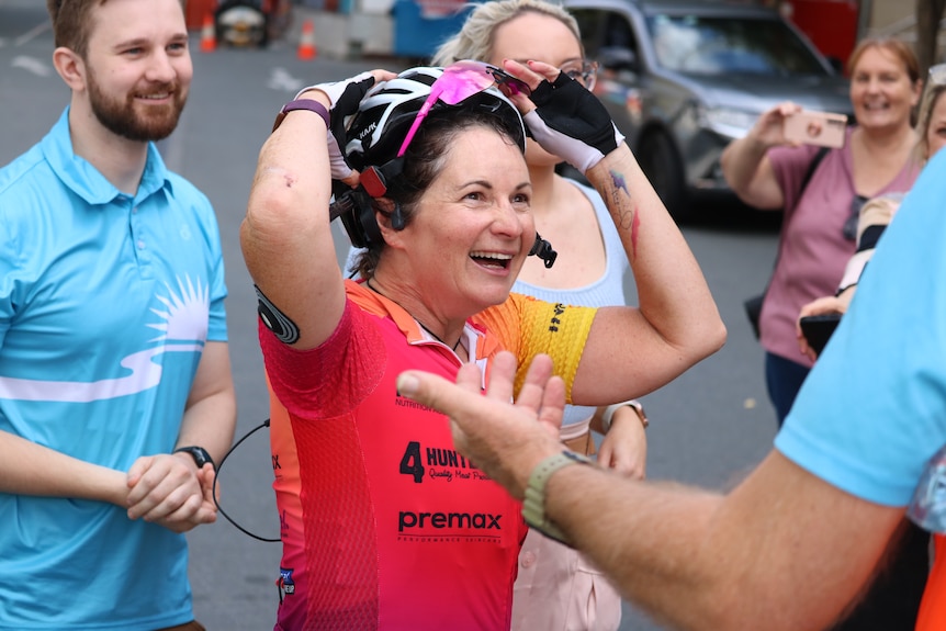 A sweaty woman in cycling gear smiles in half-shock as she is surrounded by smiling friends and family.