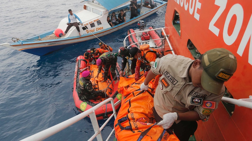 Rescuers carry body bags up stairs on a ship, with smaller boats in the background