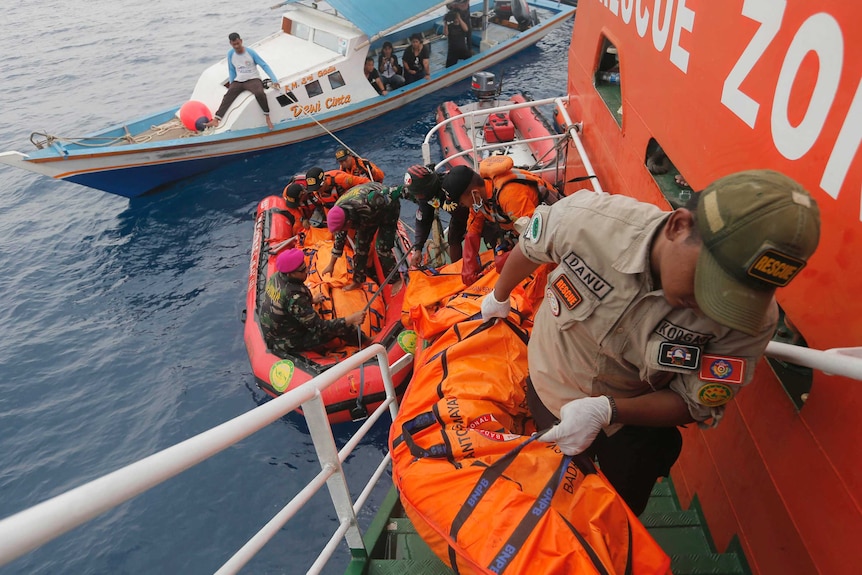 Rescuers carry body bags up stairs on a ship, with smaller boats in the background
