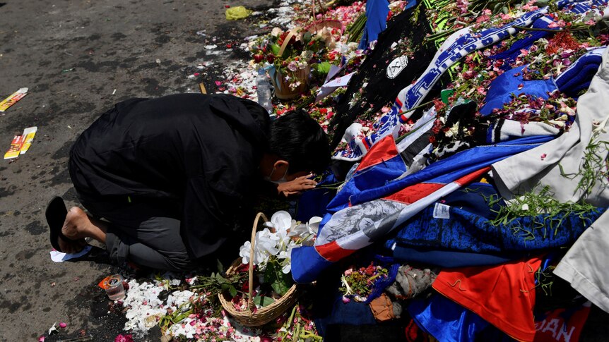 A man kneels at a memorial that is made of flowers and football merchandise apparel.