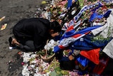 A man kneels at a memorial that is made of flowers and football merchandise apparel.