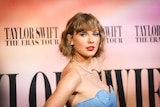 Taylor Swift dressed in a blue dress looks over her shoulder while standing in front of a sign saying Taylor Swift Eras Tour