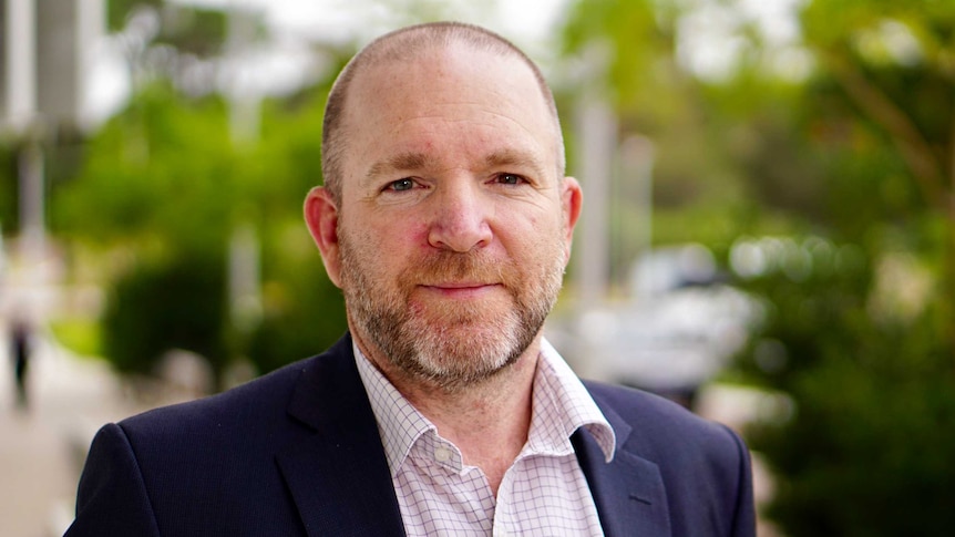 A mid shot of a man in a button-up shirt and jacket looking at the camera with greenery in the background.