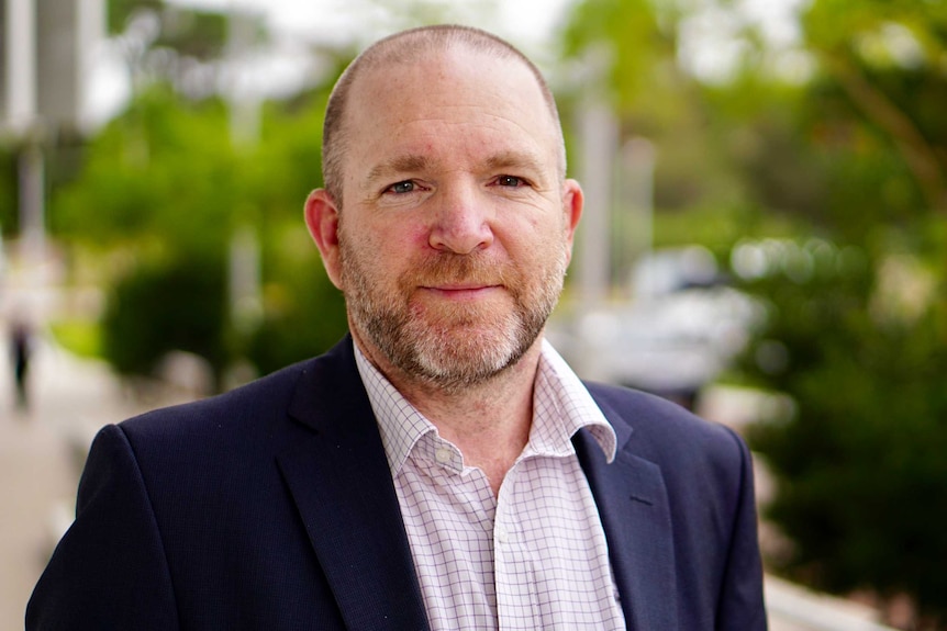 A mid shot of a man in a button-up shirt and jacket looking at the camera with greenery in the background.