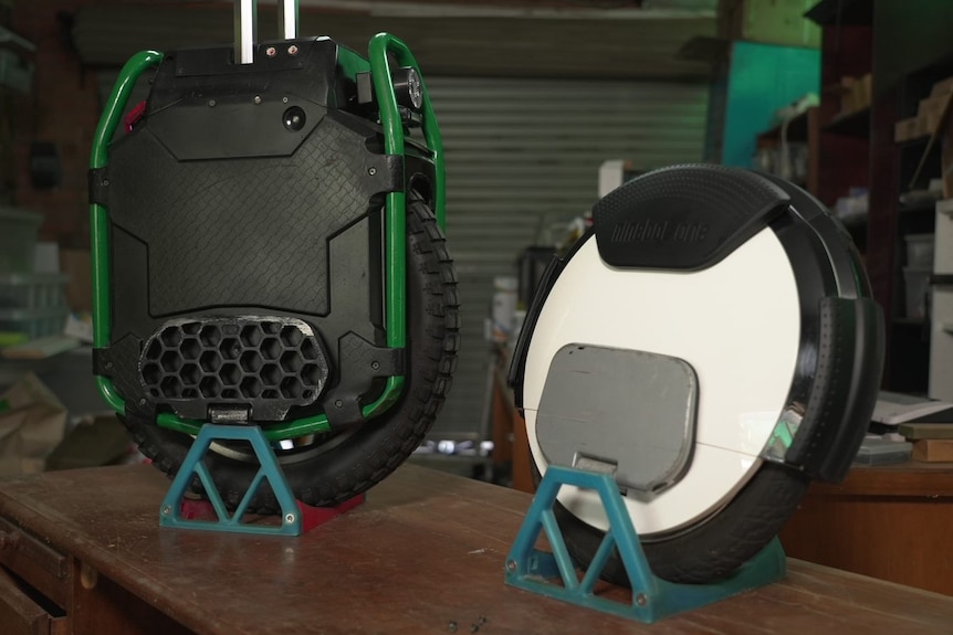 Two mobility devices on small stands in a shed. One looks like a unicycle, the other is flat and round.