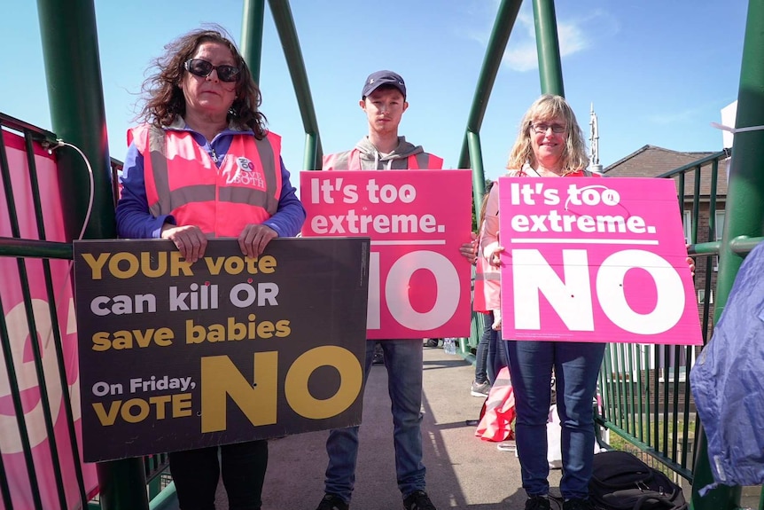 Three people hold "vote NO" signs, that say "it's too extreme".