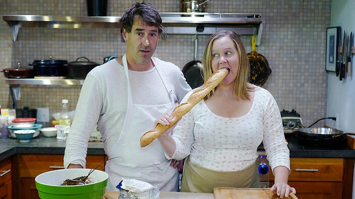 A man and woman stand in a kitchen, with the woman biting into a breadstick