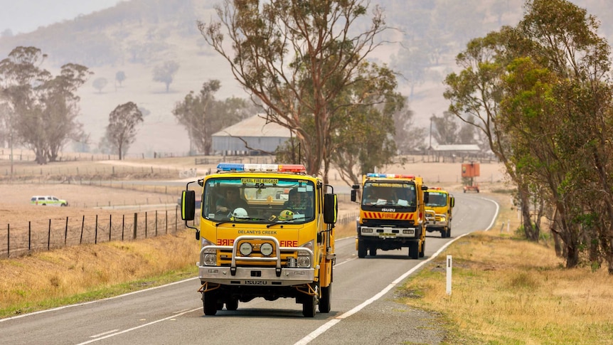 A yellow fire truck drives along a rural road through smoke.