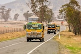 A yellow fire truck drives along a rural road through smoke.
