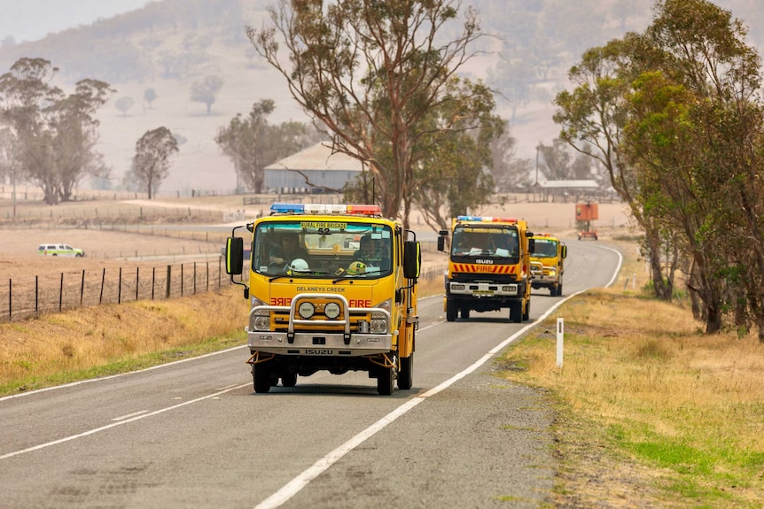 A yellow fire truck drives along a rural road through smoke.