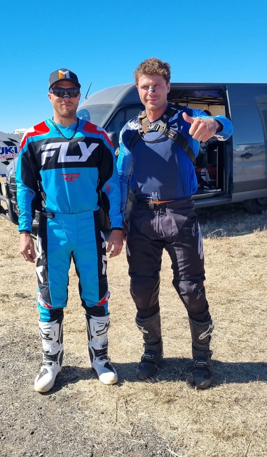 Two men standing in an outback landscape wearing motorbike riding gear, one is giving a thumbs up to the camera.