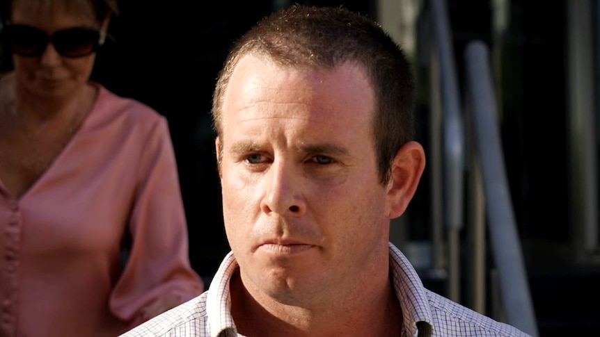 A close-up photo of a man with a solemn expression wearing a shirt with the collar open.