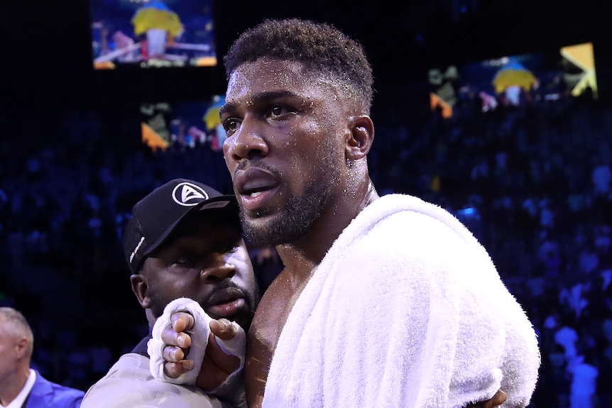 Anthony Joshua is held back by a member of his team and looks upset draped in a towel