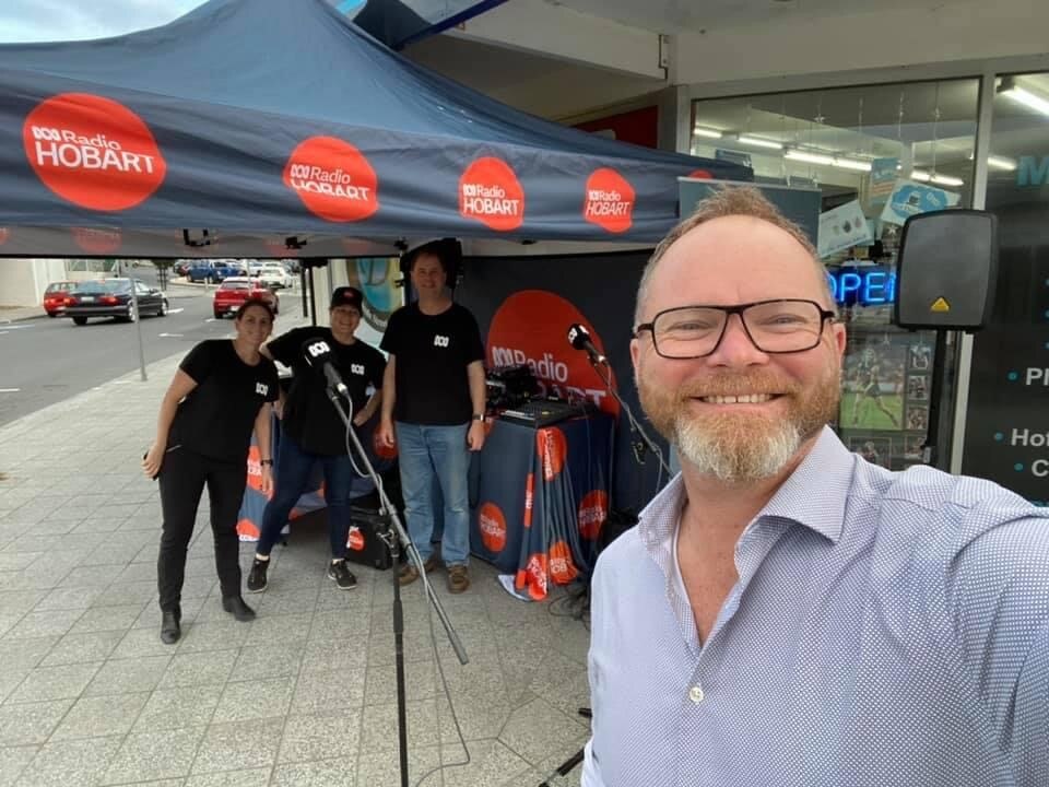 A man with glasses is taking a selfie that shows a tent with ABC Radio Hobart logo on it.