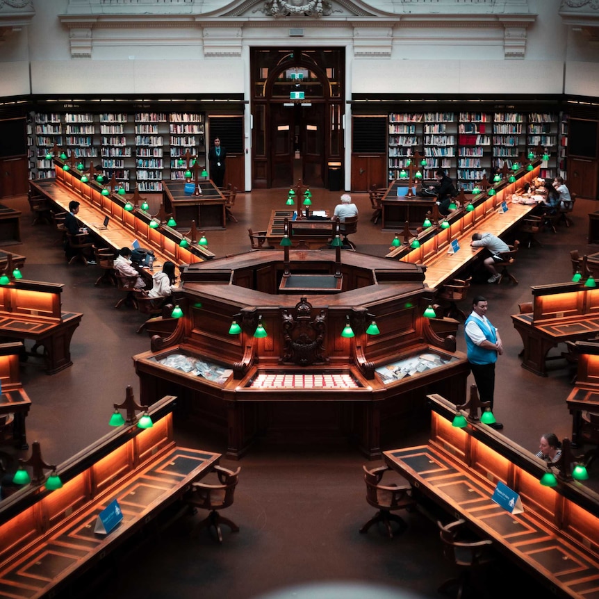 A high-angled view of an ornate orthagonal library floor, with green lamps illuminating desks radiating from the centre.