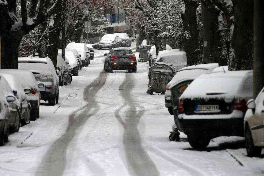 A dark Audi drives along a tree-lined street with cars parked on either side and heavy snow over all surroundings.