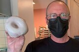 A man in a black face mask holds a white-iced doughnut with a gloved hand.